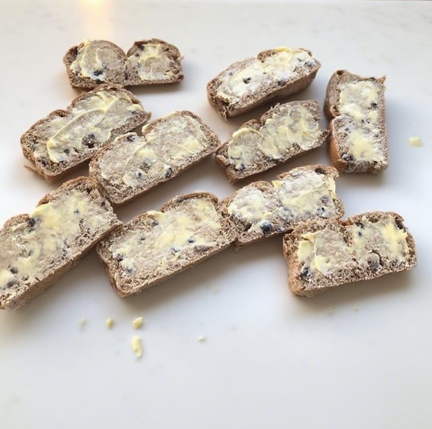  Hands buttering sliced hot cross buns on a white counter with a ceramic baking dish beside.
