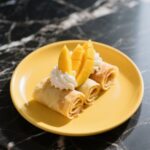 Mango crepe rolls on a yellow plate, garnished with whipped cream and mango
